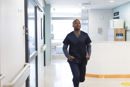 African american male doctor wearing scrubs running through corridor in hospital, copy space....