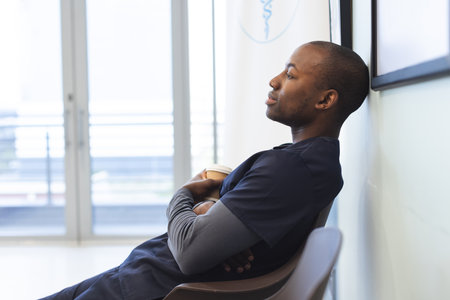 Thoughtful african american male doctor wearing scrubs sitting in waiting room in hospital....