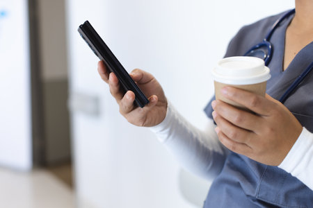 Midsection of biracial female doctor wearing scrubs using smartphone, holding takeaway coffee....