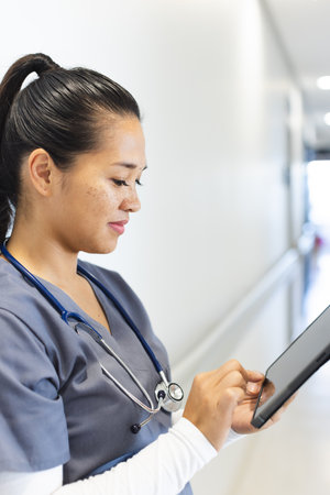 Biracial female doctor wearing scrubs using tablet in hospital corridor. Medicine, healthcare, communication, work and hospital, unaltered.の写真素材