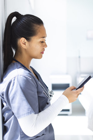 Biracial female doctor wearing scrubs using smartphon in hospital. Medicine, healthcare,...