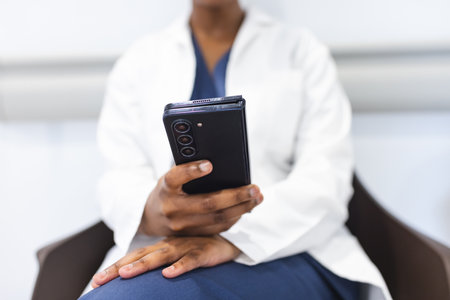 Midsection of african american female doctor using smartphone in waiting room in hospital. Medicine, healthcare, communication, work and hospital.の写真素材