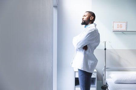 Thoughtful african american male doctor looking through window in hospital room, copy space. Medicine, healthcare, work and hospital, unaltered.の写真素材
