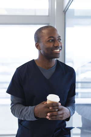 Happy african american male doctor holding takeaway coffee looking through window in hospital room....