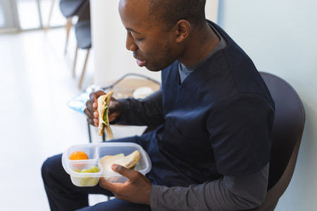 African american male doctor wearing scrubs eating takeaway food in waiting room in hospital. Medicine, healthcare, work and hospital, unaltered.の写真素材