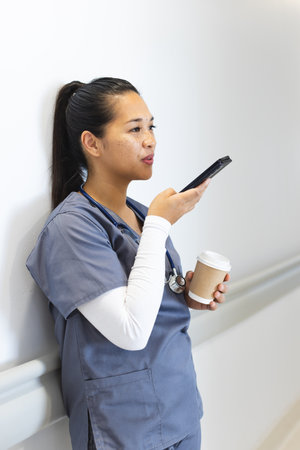 Biracial female doctor talking on smartphone, holding takeaway coffee in hospital corridor. Medicine, healthcare, communication, work and hospital, unaltered.の写真素材
