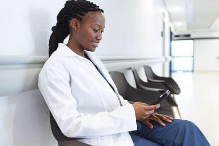 African american female doctor wearing lab coat using smartphone in hospital corridor. Medicine, healthcare, communication, work and hospital, unaltered.の写真素材