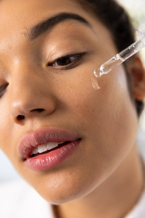 Biracial woman in bathrobe applying cosmetic on her face in sunny bathroom. Lifestyle, self care,...
