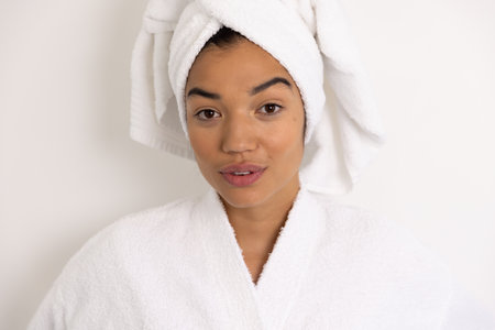 Portrait of happy biracial woman in bathrobe and with towel against white wall. Lifestyle, self care and domestic life, unaltered.の写真素材