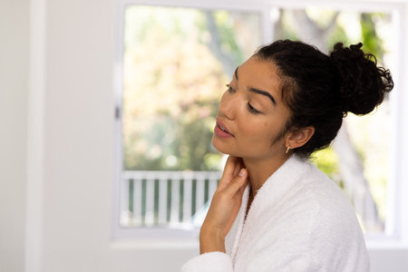 Biracial woman in bathrobe looking in mirror in sunny bathroom. Lifestyle, self care and domestic life, unaltered.の写真素材