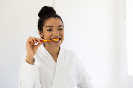 Happy biracial woman in bathrobe brushing teeth in sunny bathroom. Lifestyle, self care, hygiene and...