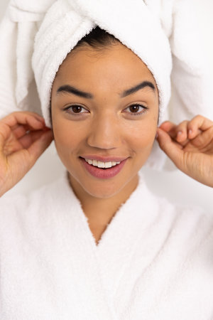 Close up portrait of happy biracial woman in bathrobe and with towel against white wall. Lifestyle, self care and domestic life, unaltered.の写真素材
