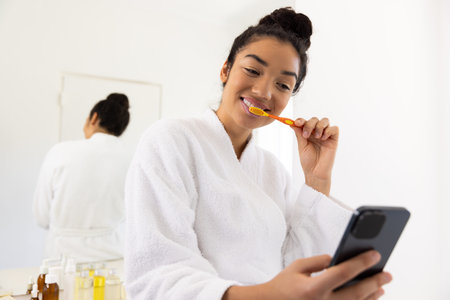 Happy biracial woman in bathrobe brushing teeth and using smartphone in sunny bathroom. Lifestyle, self care, hygiene, communication and domestic life, unaltered.の写真素材