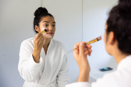 Biracial woman in bathrobe applying face mask using brush in sunny bathroom. Lifestyle, self care, beauty, skin and domestic life, unaltered.の写真素材