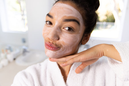 Portrait of biracial woman in bathrobe with face mask in sunny bathroom. Lifestyle, self care, beauty, skin and domestic life, unaltered.の写真素材