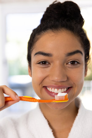 Portrait of happy biracial woman in bathrobe holding brush with tooth paste in sunny bathroom. Lifestyle, self care, beauty, skin and domestic life, unaltered.の写真素材