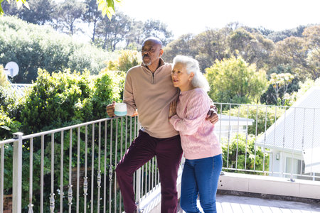 Happy diverse senior couple holding cups of coffee and embracing on sunny terrace. Lifestyle, retirement, senior lifestyle, nature, togetherness and domestic life, unaltered.の写真素材
