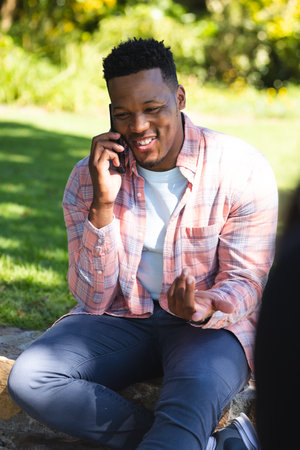 Happy african american man sitting on stairs and talking on smartphone in sunny garden, copy space. Outdoors, expression, gesticulation, technology, communication and nature, unaltered.の写真素材