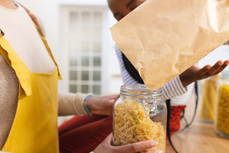 African american mother and daughter pouring pasta to glass jar in kitchen at home, copy space....