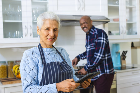 Happy diverse senior couple preparing meal using tablet in kitchen. Lifestyle, retirement, senior lifestyle, food, cooking, togetherness, communication, recipe and domestic life, unaltered.の写真素材