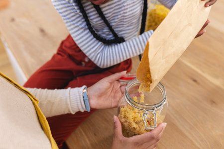 African american mother and daughter pouring pasta to glass jar in kitchen at home, copy space. Expression, togetherness, motherhood, childhood, food and drink and domestic life,の写真素材