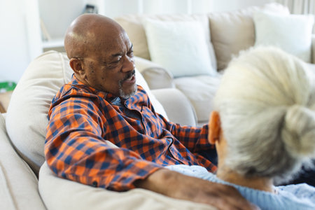 Diverse senior couple sitting on sofa, talking and embracing in sunny living room. Lifestyle, retirement, senior lifestyle, togetherness and domestic life, unaltered.の写真素材