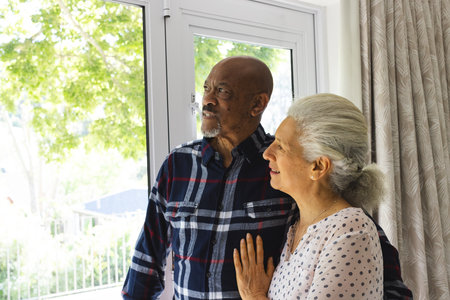 Diverse senior couple embracing and looking out window at sunny home. Lifestyle, retirement, senior...
