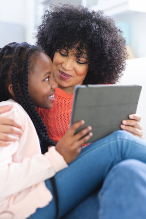 Happy african american mother and daughter embracing on couch and using tablet at home, copy space. Motherhood, childhood, togetherness, technology, communication and domestic life, unaltered.の写真素材