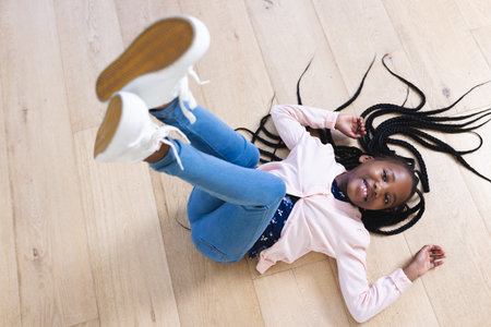 Happy african american girl lying on floor with legs in air at home, copy space. Childhood, expression, gesticulation and domestic life, unaltered.の写真素材