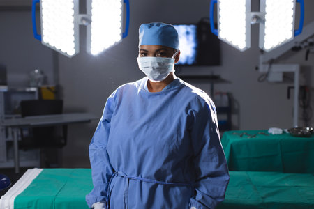 Portrait of biracial female surgeon wearing surgical gown and face mask in operating theatre. Medicine, healthcare, surgery, work and hospital, unaltered.の写真素材