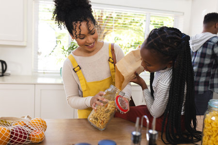 Happy african american mother and daughter pouring pasta to glass jar in kitchen at home, copy space. Expression, togetherness, motherhood, childhood, food and drink and domestic life,の写真素材
