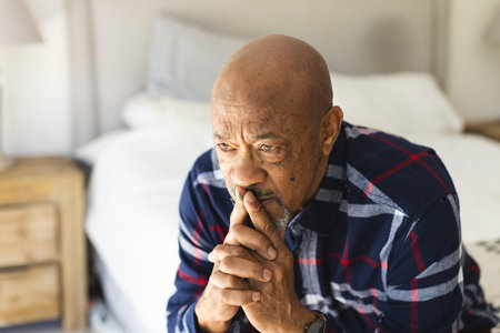 Thoughtful african american senior man sitting on bed in sunny bedroom. Lifestyle, retirement, senior lifestyle and domestic life, unaltered.の写真素材