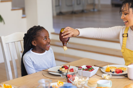 Happy african american mother pouring maple syrup on fruits in dining room at home, copy space. Togetherness, motherhood, childhood, food and drink and domestic life, unaltered.の写真素材