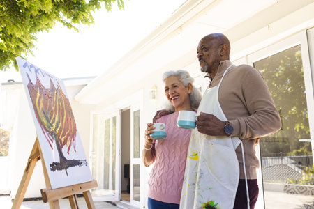 Happy diverse senior couple drinking coffee, embracing and looking at painting on sunny terrace. Lifestyle, retirement, senior lifestyle, nature, creativity, togetherness and domestic life, unaltered.の写真素材