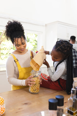 Happy african american mother and daughter pouring pasta to glass jar in kitchen at home, copy...
