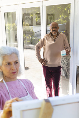 Diverse senior woman painting on canvas and senior man drinking coffee on sunny terrace. Lifestyle, retirement, senior lifestyle, nature, creativity, togetherness and domestic life, unaltered.の写真素材