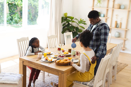 Happy african american man pouring tea to cup at breakfast in morning at home, copy space. Togetherness, parenthood, childhood, food and drink, domestic life and healthy lifestyle, unaltered.の写真素材