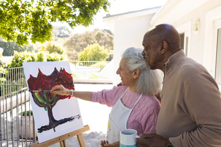Happy diverse senior woman painting on canvas and senior man embracing her on sunny terrace. Lifestyle, retirement, senior lifestyle, nature, creativity, togetherness and domestic life, unaltered.の写真素材