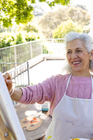 Happy caucasian senior woman painting on canvas on sunny terrace. Lifestyle, retirement, senior lifestyle, nature, creativity and domestic life, unaltered.の写真素材