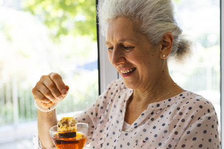 Happy caucasian senior woman drinking tea in window at sunny home. Lifestyle, retirement, senior lifestyle and domestic life, unaltered.の写真素材