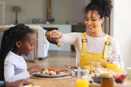 Happy african american mother pouring maple syrup on fruits in dining room at home, copy space. Togetherness, motherhood, childhood, food and drink and domestic life, unaltered.の写真素材