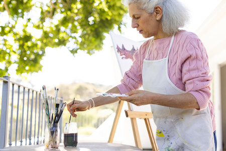Caucasian senior woman painting on canvas on sunny terrace. Lifestyle, retirement, senior lifestyle, nature, creativity and domestic life, unaltered.の写真素材