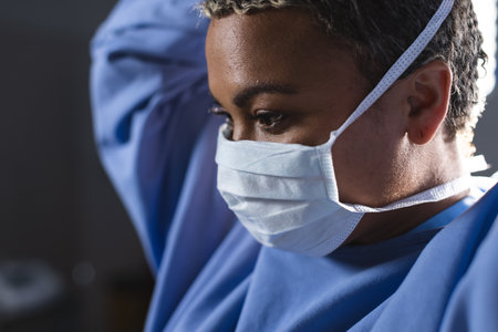 Biracial female surgeon wearing surgical gown and face mask in operating theatre. Medicine,...