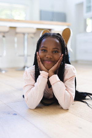 Happy african american girl lying on floor with hands on face at home, copy space. Childhood, expression, gesticulation and domestic life, unaltered.の写真素材