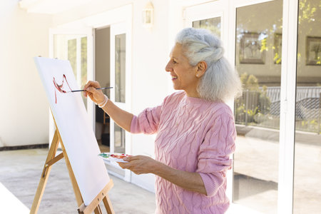 Happy caucasian senior woman painting on canvas on sunny terrace. Lifestyle, retirement, senior lifestyle, creativity and domestic life, unaltered.の写真素材