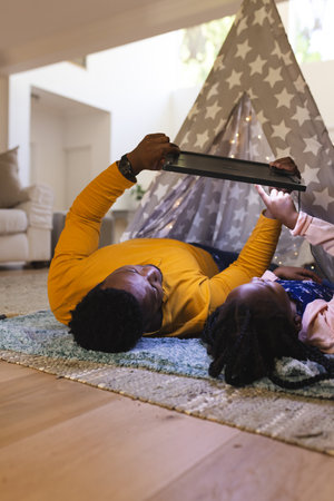 Happy african american father and daughter lying by teepee with tablet at home, copy space....