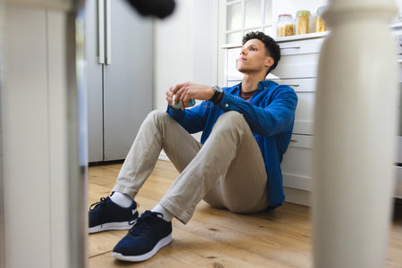Concerned biracial man sitting on floor and holding tea cup in kitchen at home, copy space....
