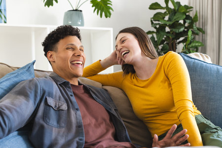 Happy diverse couple sitting on couch and laughing in living room at home, copy space. Expression, love, togetherness and domestic life unaltered.の写真素材