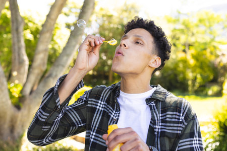 Biracial man blowing soap bubbles in sunny garden, copy space. Outdoors, expression, leisure, fun and nature unaltered.の写真素材