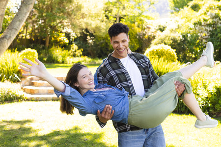 Happy diverse man holding woman in arms in air in sunny garden, copy space. Outdoors, expression, love, togetherness, fun and nature unaltered.の写真素材